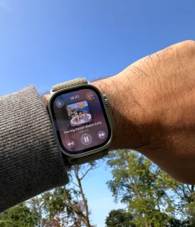 Poignet d'une personne portant une montre connectée affichant une application de lecture audio avec un fond d'arbres et un ciel bleu.