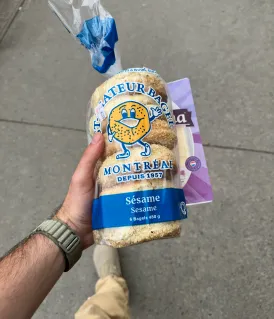 Main tenant un paquet de pain de blé entier au sésame de la marque Fratelli Montreux avec un sac plastique bleu attaché en haut.