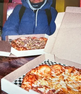 Person wearing a blue jacket holding an open pizza box with a pizza inside, with another open pizza box on a table in the foreground.
